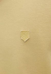 Geltona tekstilė su tekstūruotu paviršiumi, kuriame yra mažas išsiuvinėtas skydo logotipas šiek tiek šviesesnėje geltonoje spalvoje.