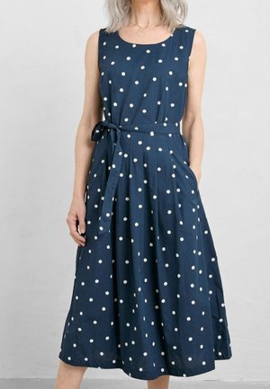 Mujer con vestido midi azul marino sin mangas con lunares blancos y cinturón atado en la cintura, de pie con una mano en el bolsillo.