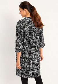 Vrouw met een lange, bruine paardenstaart, gekleed in een zwart-wit patroon knie-lengte jurk en zwarte leggings, staand met gesloten ogen, met de rug naar voren gekeerd.