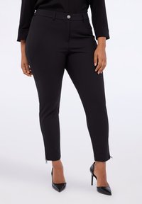 Pantalones ajustados negros hechos de tela suave, con cremallera delantera y cierre de botón, piernas entalladas y cremalleras en los tobillos para mayor ajustabilidad.