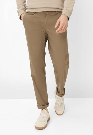 Uomo che cammina indossando pantaloni beige risvoltati, felpa beige chiaro e sneakers color crema con suole in gomma.