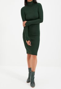 Abito a coste di maglia verde scuro con collo alto e maniche lunghe, silhouette aderente, abbinato a stivaletti verdi alla caviglia.