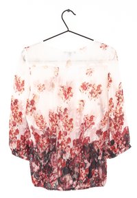 Blouse transparente, motif floral dans des nuances de rouge, rose et noir. Manches longues, ourlet froncé, poignets boutonnés, suspendue à un crochet noir.