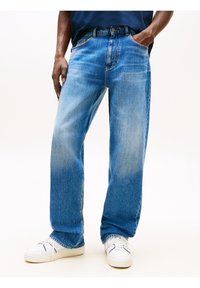 Homme portant un jean large bleu clair, des baskets blanches et une chemise bleu marine, debout les mains dans les poches.