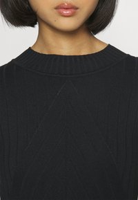 Femme aux cheveux courts et foncés, lèvres brillantes, portant un pull noir en maille côtelée avec un motif chevron sur la poitrine.