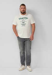 Cremfarbene T-Shirt mit dunkelgrünem Grafikdruck, kombiniert mit grauen Jeans und hellen Sneakers. Das Shirt hat kurze Ärmel und eine lockere Passform.