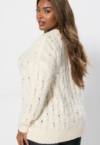 Neule vaalean kermanvärinen neule, jossa on tekstuurinen langankäyttökuvio, leveät hihat ja ribbireuna. Sisältää avoimen neuleen suunnittelun hengittävyyden parantamiseksi.