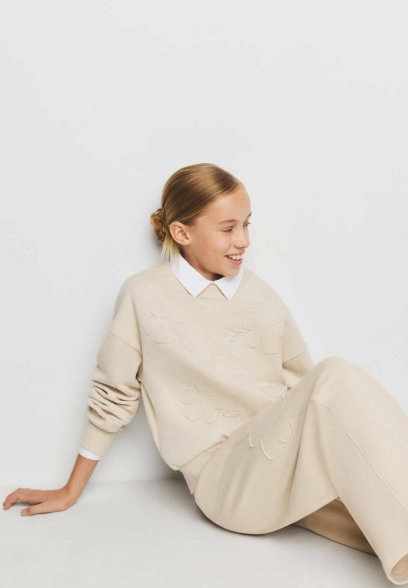 Fille aux cheveux châtain clair en chignon, vêtue d'un pull beige à motif floral et d'une jupe assortie, souriante et assise contre un mur clair.