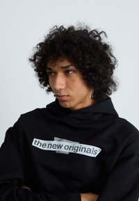 Fleeced preto com um grande texto gráfico branco "os novos originais" com efeito de fita transparente. O modelo tem cabelo encaracolado e um piercing no septo.