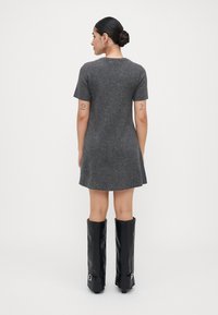 Donna con capelli scuri raccolti in un chignon, che indossa un vestito grigio a maniche corte e stivali neri al ginocchio, in piedi rivolta di spalle contro uno sfondo semplice.