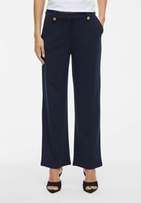 Vrouw in marineblauwe wijde broek met gouden knopen op de taille, gecombineerd met zwarte open teen hakken, staand met handen in de zakken.