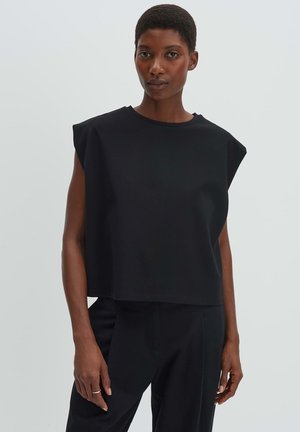 Vrouw met kort haar, gekleed in een mouwloos zwart topje met gewatteerde schouders en een zwarte broek, tegen een effen lichte achtergrond.