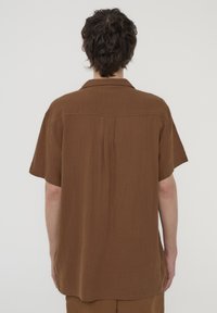 Una camicia marrone a maniche corte realizzata in tessuto testurizzato con colletto; presenta un yoke e spacchi laterali all'orlo per facilitare i movimenti.