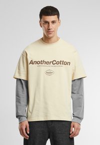 T-shirt en coton beige superposé sur une chemise grise à manches longues. présente un logo en texte marron "AnotherCotton" et un slogan supplémentaire en dessous.