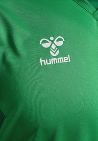 Groen sportshirt gemaakt van textuurstof met een wit logo en de tekst "hummel" aan de linkerkant. V-halsontwerp met contrasterende stiksels.