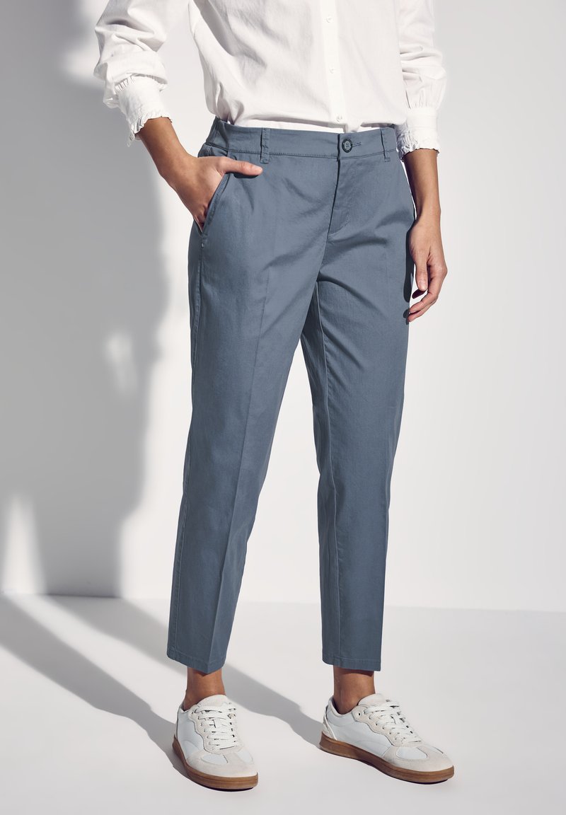 Pantaloni su misura di un azzurro chiaro, dal tessuto liscio, con chiusura a bottone e tasche laterali, abbinati a sneaker bianche.