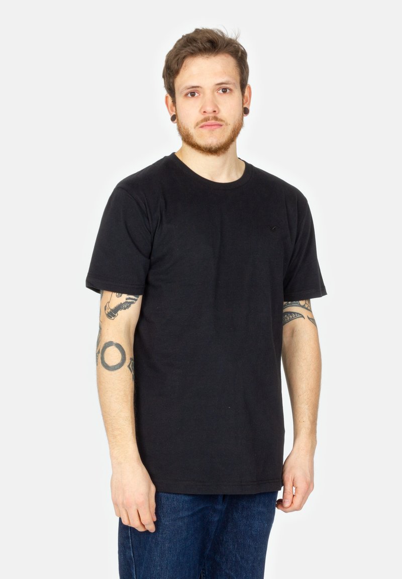 Schwarzes Baumwoll-T-Shirt mit kurzen Ärmeln, rundem Halsausschnitt und subtiler Logodetailierung auf der Brust. Kombiniert mit blauen Jeans.