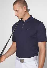 Navy polo shirt with short sleeves, three-button placket, and a small white logo. Paired with a white belt and holding a golf club.