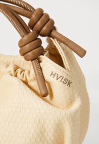 Rund beige handväska med vävd textur. Bruna läderdragsnören och detaljer. Varumärket "HVISK" broderat på sidan.