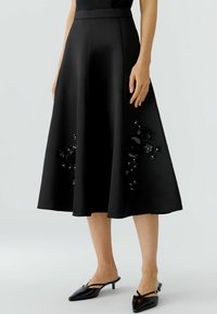 Jupe midi noire avec des ornements en sequins floraux sur les côtés, portée avec des escarpins à bout pointu noirs sur un fond clair.