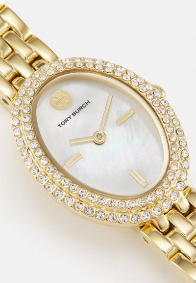 Gouden Tory Burch polshorloge met ovale parelmoeren wijzerplaat, kristal bezette lunette, en gouden uurmarkeringen en wijzers op een schakelarmband.