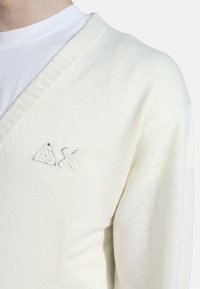 Maglione a maglia di colore crema chiaro con scollatura a V, caratterizzato da un colletto a coste texturizzato e un logo ricamato a forma di triangolo e croce.