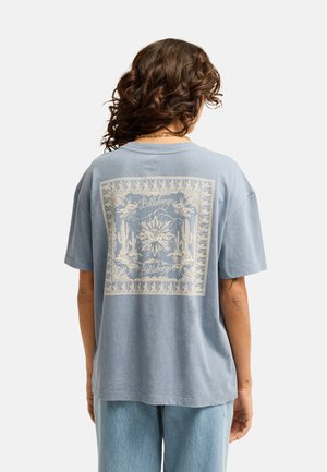 Kobieta o kręconych brązowych włosach nosi wyblakły niebieski T-shirt z beżową grafiką o tematyce pustynnej oraz jasnoniebieskie dżinsy, widziana od tyłu.