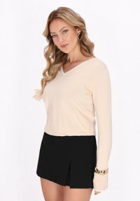 Top cu mânecă lungă, de tip V, în nuanța bej deschis, cu textură netedă; asortat cu o fustă mini plisată neagră; brățară cu accente aurii pe încheietura mâinii.
