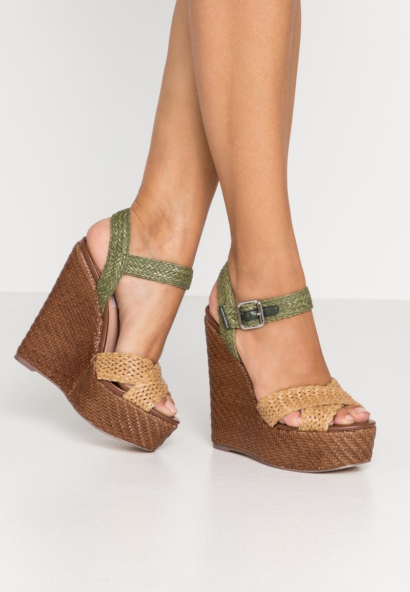 Pieds portant des sandales compensées marron en tissage avec des brides de cheville vertes tressées et des boucles sur un sol blanc.