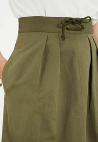 Jupe vert olive en tissu texturé, dotée de plis, de poches latérales et d'une taille à cordon de serrage pour un ajustement réglable.