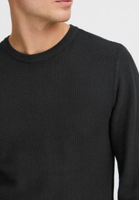 Svart teksturert strikkegenser med ribbestrikket rund hals. Tøyet har et distinkt kryssmønster som gir ekstra dybde og detaljer.