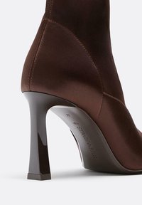 Brauner, knöchelhoher Stiefel mit hohen Absätzen aus glattem Obermaterial, sichtbaren Nähten und einem glänzenden, verjüngten Blockabsatz auf weißem Hintergrund.