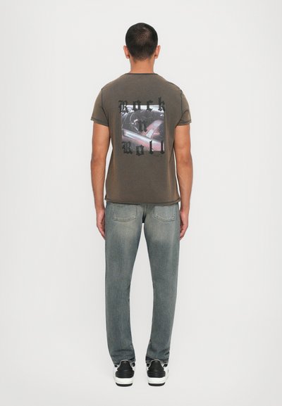 Olive green t-shirt featuring a graphic print with "Rock n Roll" text. Paired with light gray jeans and black and white sneakers.