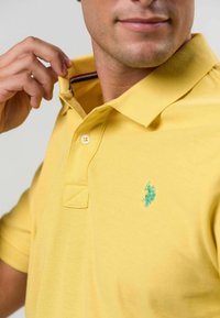 Žluté polo tričko s texturovanou tkaninou, vybavené dvouknoflíkovou légu a vyšívaným tyrkysovým logem na hrudi.