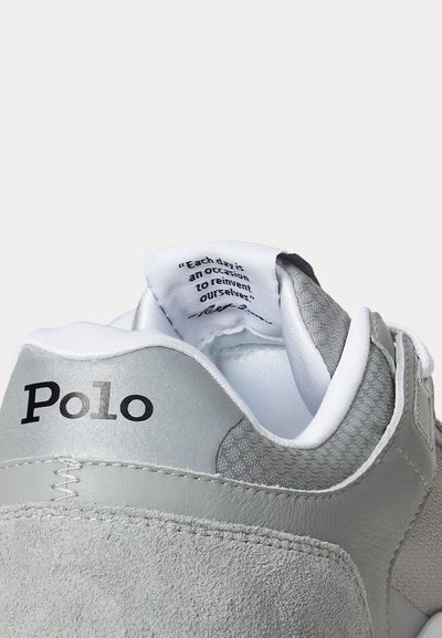 Baskets Polo grises et blanches affichant le nom de la marque sur le talon et une étiquette sur la languette avec la citation, "Each day is an occasion to reinvent ourselves."