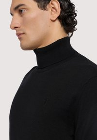 Svart polotröja i mjukt material, kroppsnära design, ribbad krage, har en liten logotyp på bröstet, sidovy av modellen.