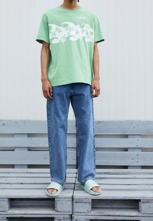 Person iført en lys grøn grafisk t-shirt, blå jeans og matchende lys grønne slidesandaler stående på træpaller.