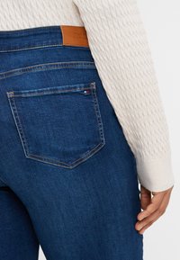 Dark blue denim jeans with a leather label on the waistband, featuring a back pocket with a small logo accent. Textured beige sweater sleeve visible.