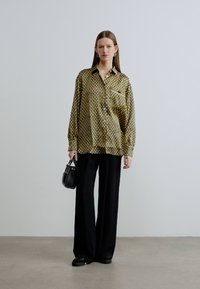 Rakstaina pogājama krekla ar garām piedurknēm olīvu zaļā un krēmkrāsā, brīvs piegriezums, apvienots ar melnām plašām biksēm. Tur rokās nelielu melnu somiņu.