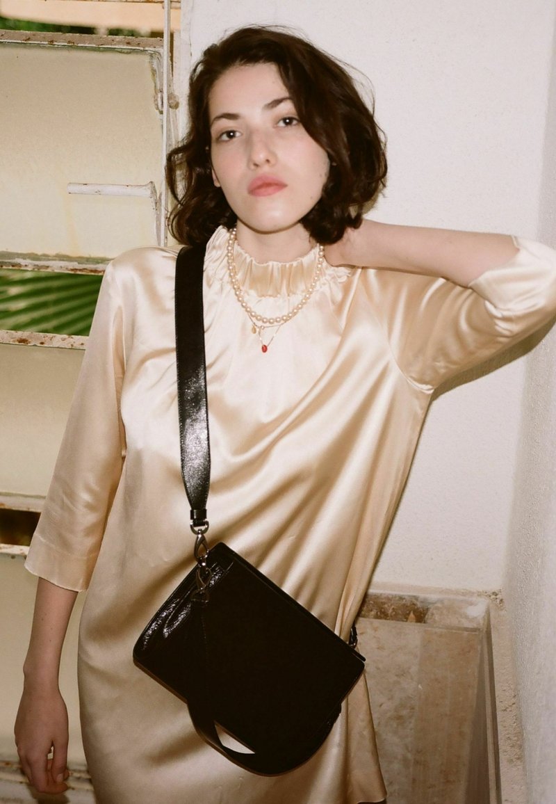 Svart läder crossbody-väska med en glänsande yta, rektangulär form och justerbar rem, kombinerad med en satinlängd klänning i krämfärg och pärlhalsband.