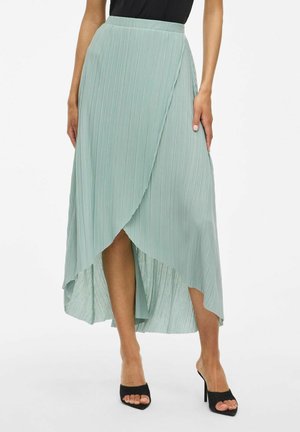 Lichtgroene, geplooide maxi-rok met een asymmetrische zoom; zachte stof benadrukt de draping; gecombineerd met zwarte open teen hakken.