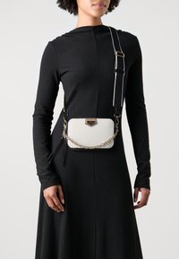 Biała skórzana torba na ramię z złotym łańcuszkiem, z fakturowanym wykończeniem i złotym akcentem metalowym.