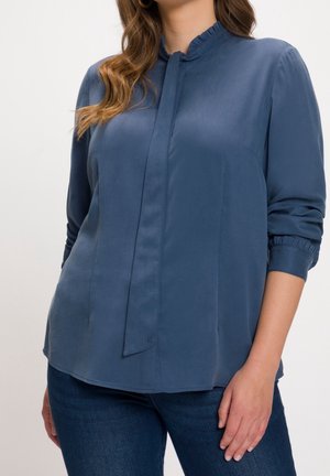 Frau trägt eine langärmlige blaue Bluse mit gerafftem Kragen und Bindendetail, kombiniert mit dunklen blauen Jeans, stehend vor einem schlichten Hintergrund.