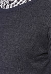 Maglione grigio scuro a girocollo con una finitura testurizzata, caratterizzato da un colletto a coste scuro e un colletto di camicia a quadri visibile sotto.