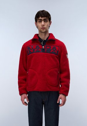 Jonge man met een rode fleecejas met "NAPA" in marineblauwe letters op de borst en een donkere broek, staand tegen een effen lichte achtergrond.