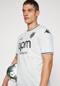 Witte voetbalshirt met korte mouwen en golfpatroon, zwarte accenten en logo; gecombineerd met een zwart-wit gestippelde voetbal.