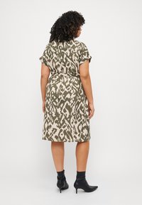 Knee-length, leopard print dress in olive green and beige, featuring short sleeves, a belted waist, and worn with black ankle boots.
