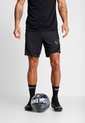 Muž ve černých krátkých šortkách Nike, černých ponožkách Nike a kopačkách stojí s černobílým fotbalovým míčem Nike F.C. u nohou.