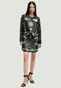 Robe en velours noir à manches longues ornée de broderies célestes argentées, comprenant des étoiles et des motifs de soleil, accompagnée de talons noirs à bride de cheville.