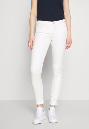Jeans skinny blancs avec une texture lisse et extensible, dotés d'un design à cinq poches, accompagnés de baskets blanches et d'un haut bleu marine.